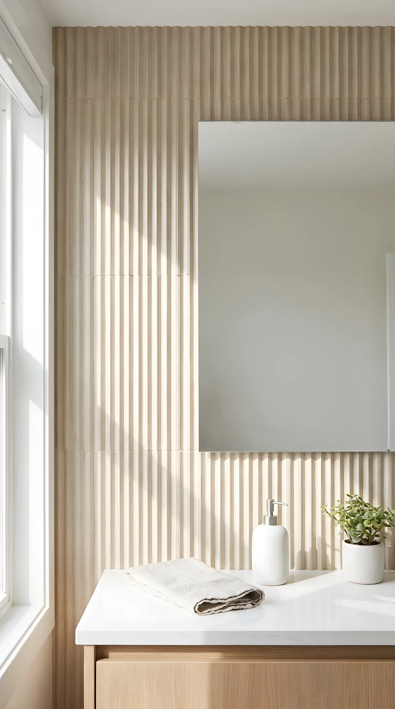 Real-life photograph of a bathroom vanity with a single fluted ceramic tile panel in soft beige behind the mirror, vertical grooves catching natural light and shadow, white countertop with neatly arranged soap dispenser, bright natural light, neatly made, organized, clean, stock photo style, no illustration.