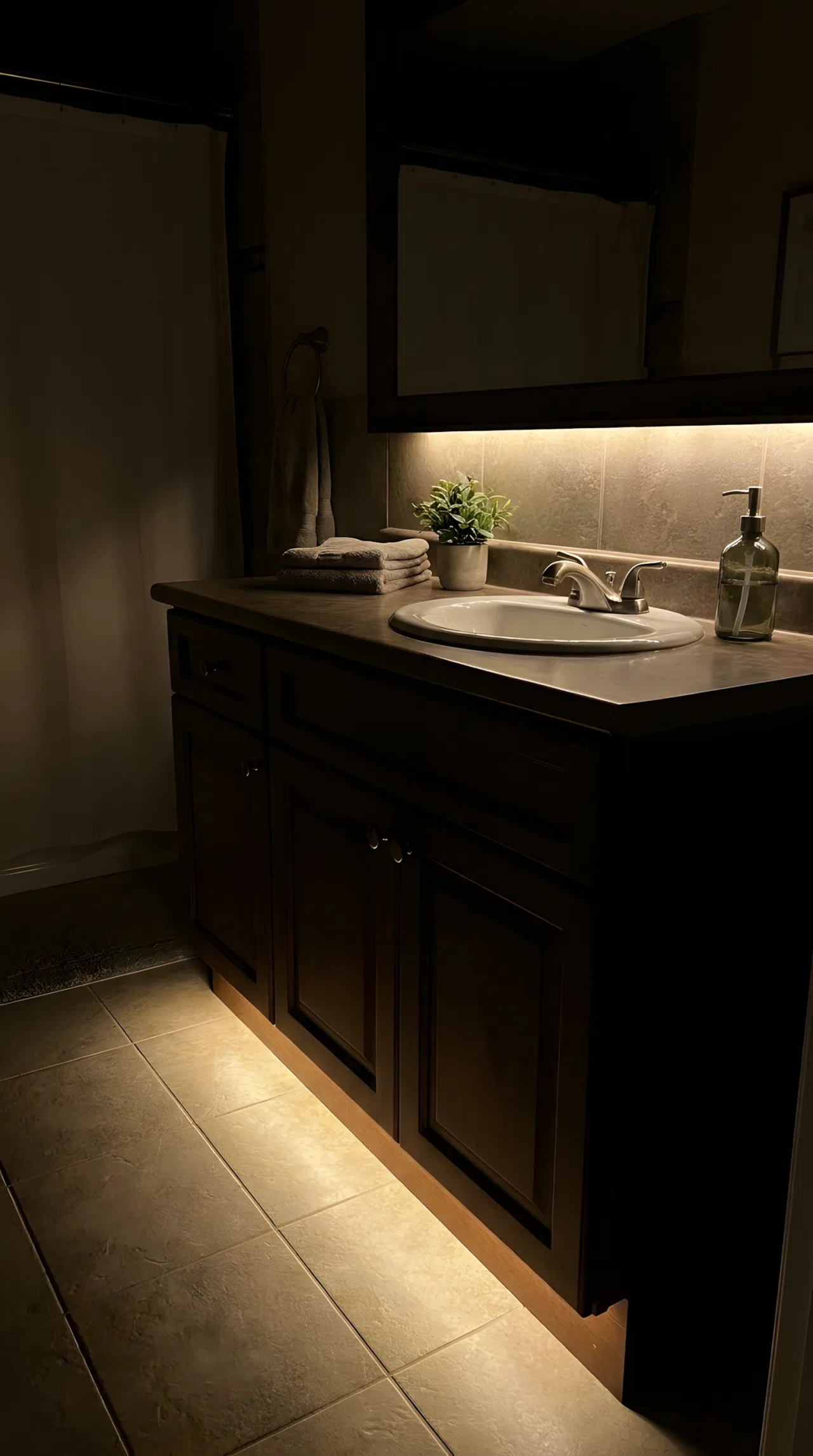 Real-life photograph of a bathroom vanity at night with a hidden LED light strip tucked into the toe-kick, soft warm light glowing upward across the lower half of a textured matte tile backsplash, rest of the room dim, cozy realistic ambiance, clean organized counter, photographic stock photo style.