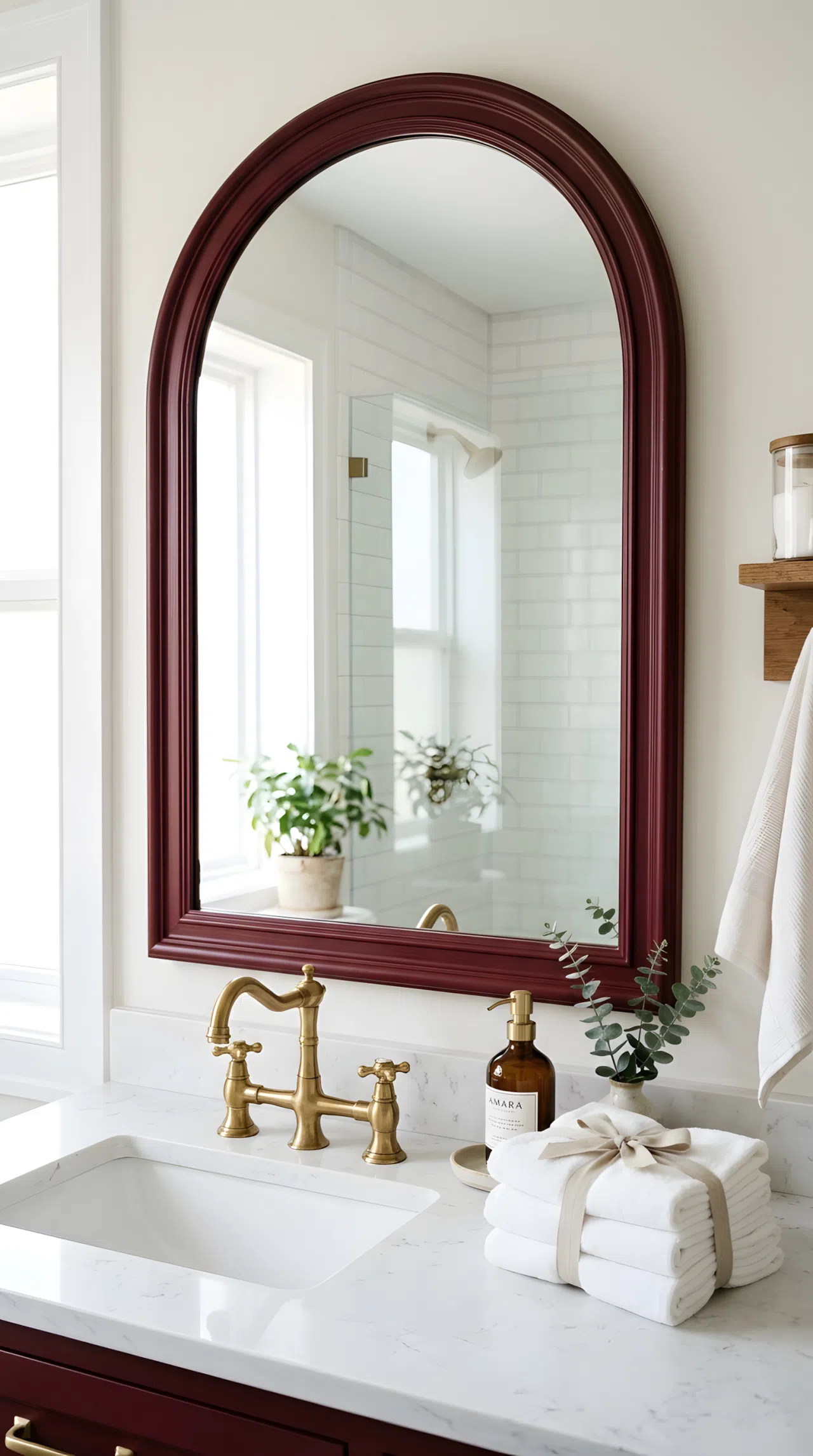 Real-life bathroom with a burgundy arched mirror frame (RGB 96,34,42), white stone vanity top, brass faucet, neatly arranged towels beautifully folded beside the sink, bright natural light reflecting off the mirror and highlighting the frame's rich color, clean organized styling, photography style.