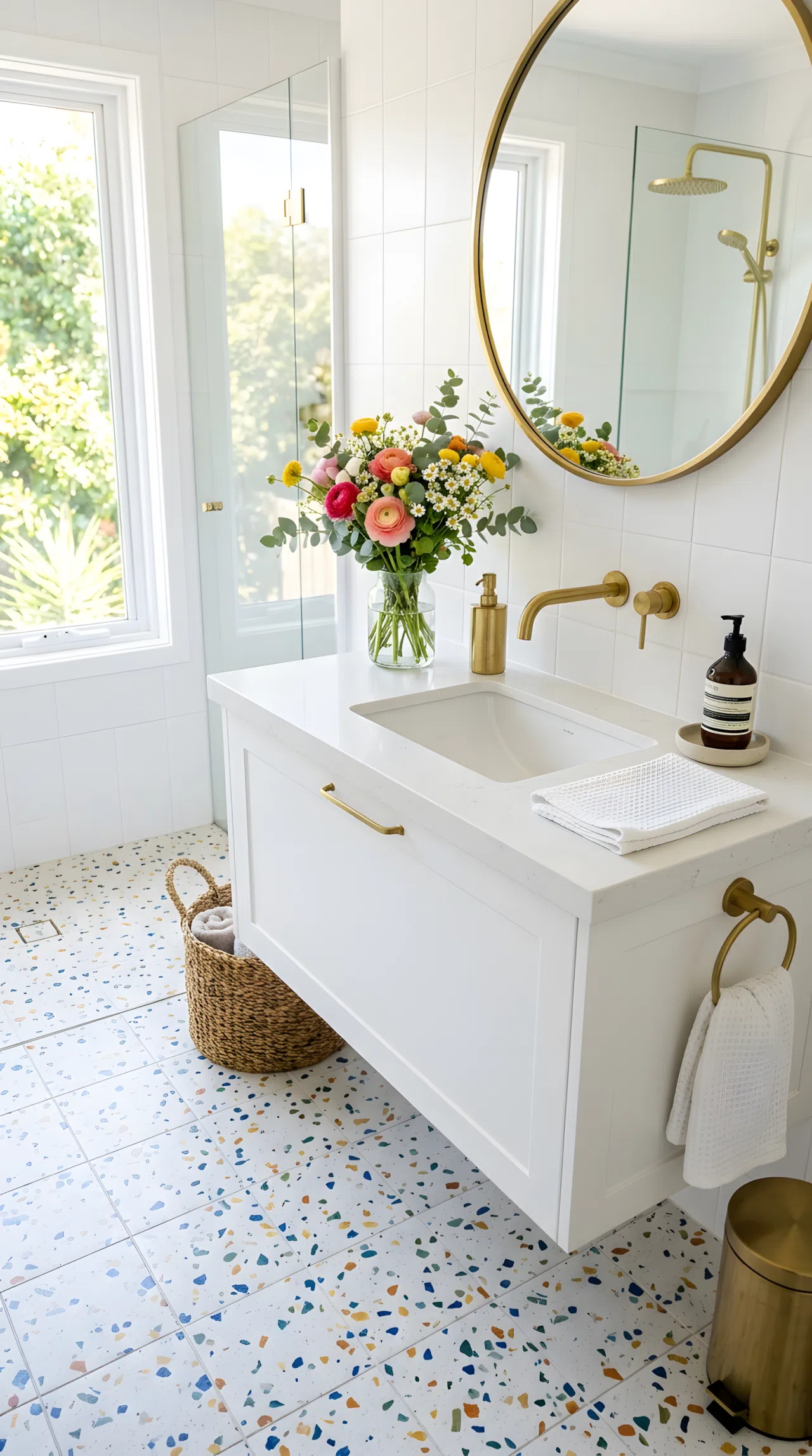 Real-life bathroom photo with white and multicolor speckled terrazzo floor tile, brass fixtures, neatly arranged white vanity, beautifully organized fresh flowers in a small vase, bright natural light, vibrant playful color flecks, clean and tidy, photographic style