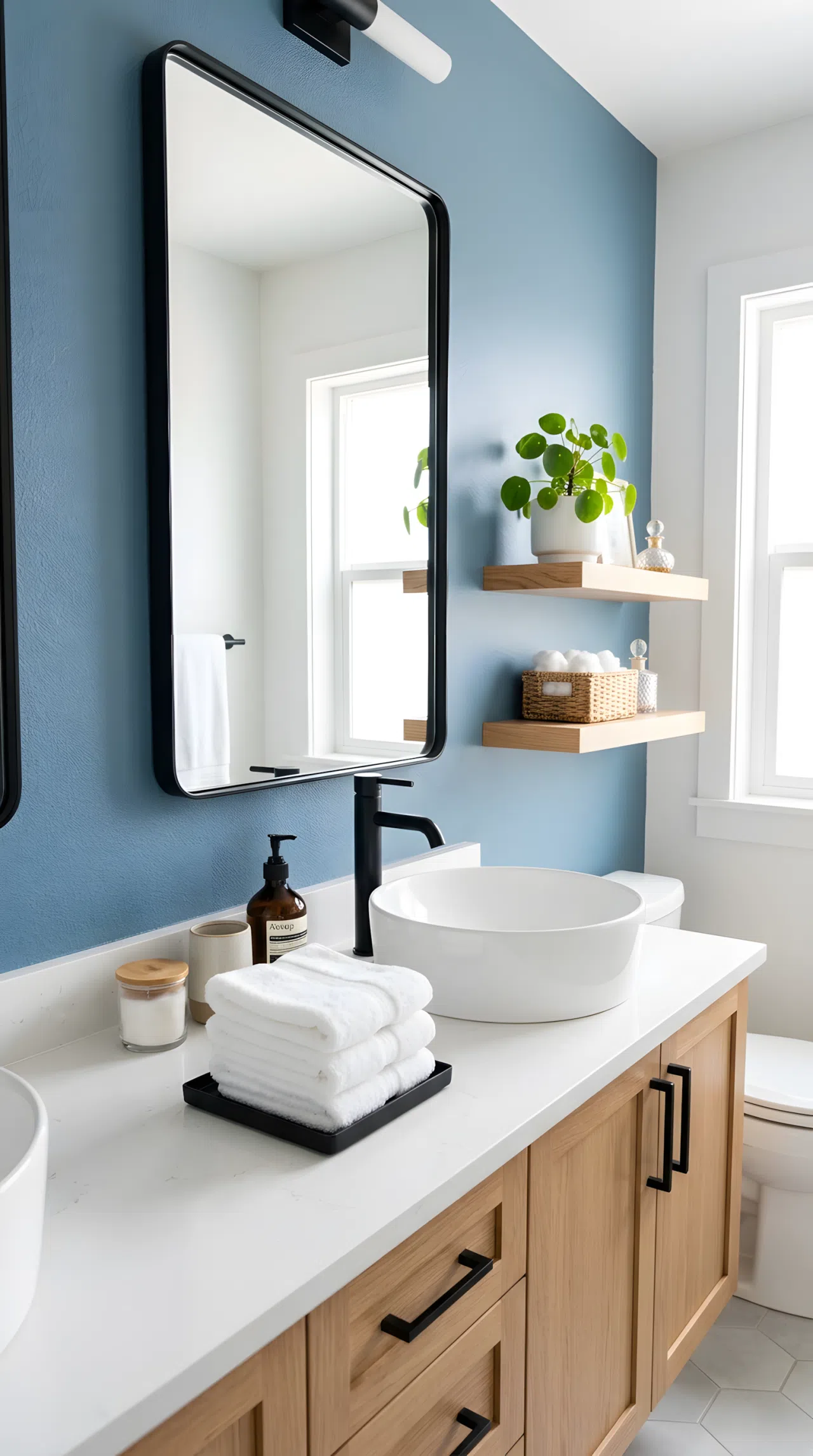 A real-life bathroom photo of a dusty blue accent wall (RGB 122,148,165) behind a matte black-framed mirror and black faucet, neatly made vanity, beautifully arranged white towels, organized shelf with one small green plant, clean, bright natural light, vibrant color contrast, photographic stock-photo style.