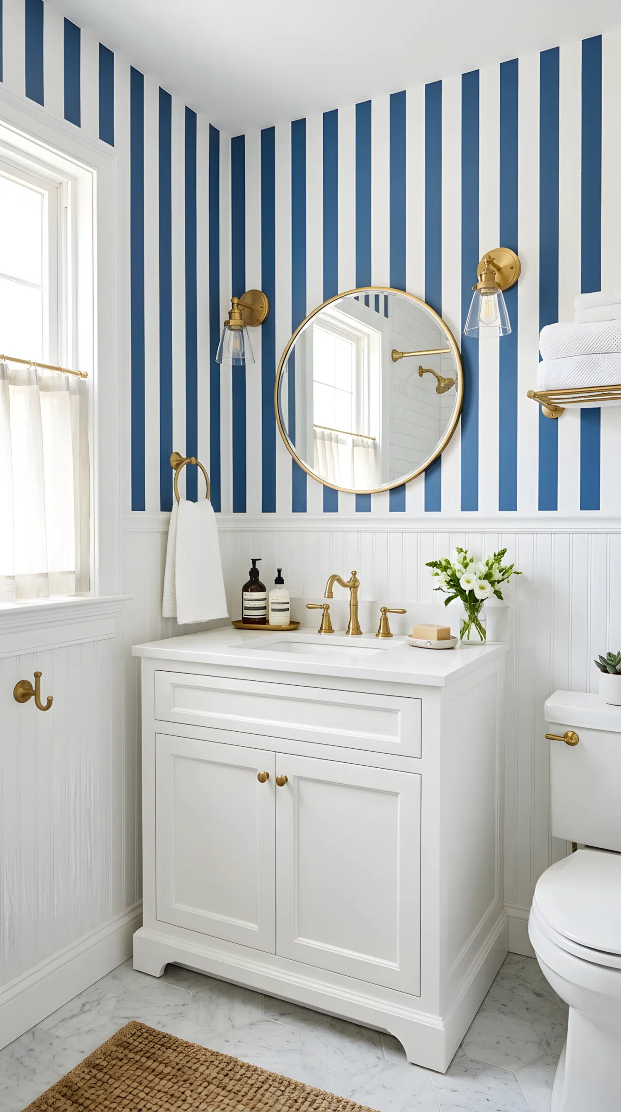 A real-life bathroom photo of blue and white striped wallpaper (RGB 58,90,140) with white wainscoting below, neatly made vanity, beautifully arranged brass fixtures, organized, clean, minimal decor, bright natural light, vibrant color, photographic stock-photo style.