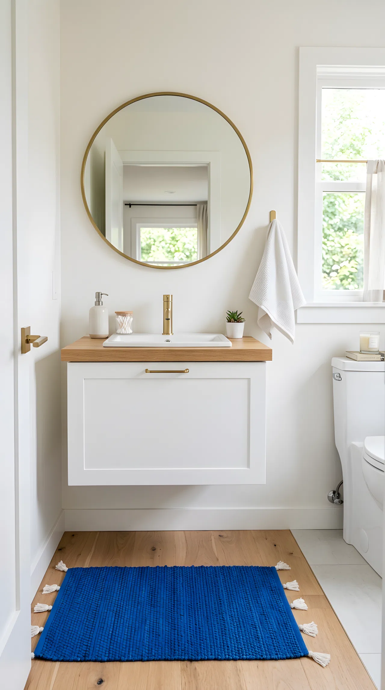 Real-life stock photo of a small guest bathroom with a white wall-mounted floating vanity, single slim drawer, round mirror above, exposed clean flooring beneath, neatly made hand towel on a hook, organized countertop, bright natural light from a window, vibrant blue bath mat accent, Pinterest-ready aesthetic, photographed like an interior design magazine, no illustration, no 3D render.